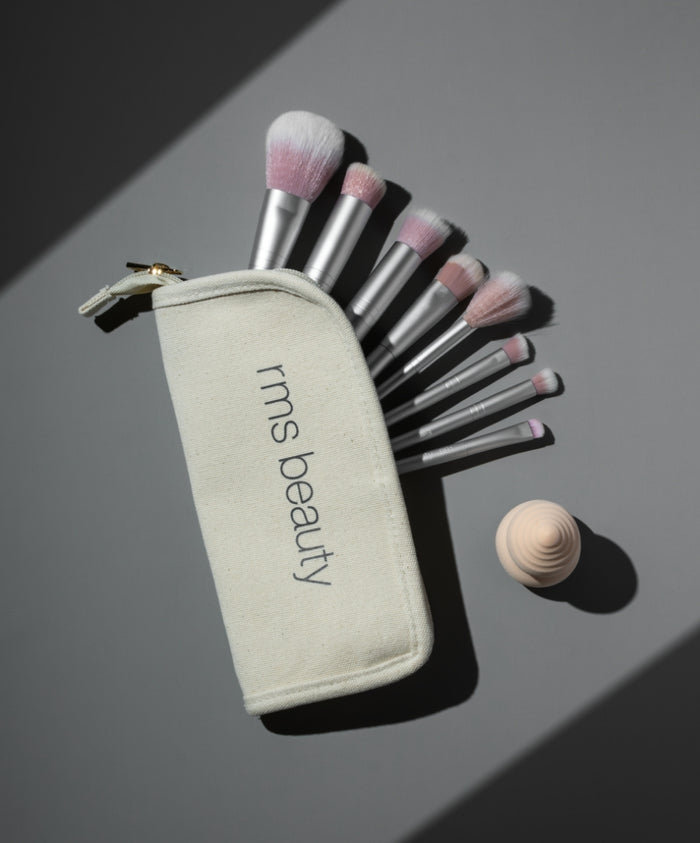 The Artist Toolkit featuring a set of soft-bristled makeup brushes with pink tips arranged alongside a natural fabric rms beauty pouch and a wooden sculpted tool, displayed on a gray surface.