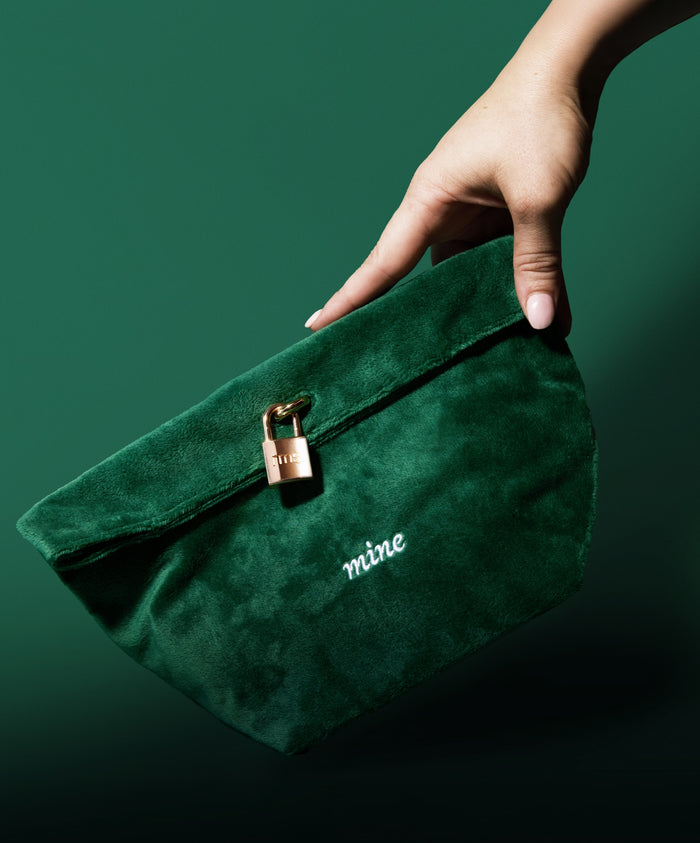 Hand holding a dark green velvet pouch with a small gold lock and the word "mine" embroidered, part of The Chameleon 2-Piece Set, emphasizing clean beauty and sustainable packaging.