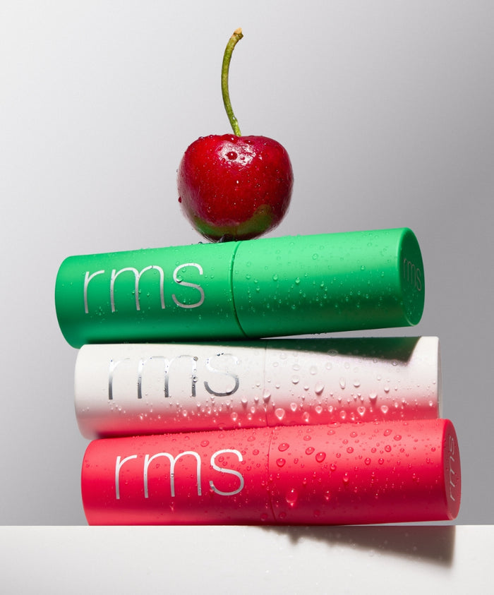 The Mini Lip Oil Trio consists of three sleek tubes in green, white, and pink, each with dewy droplets, stacked with a fresh cherry on top, highlighting clean beauty and natural ingredients.