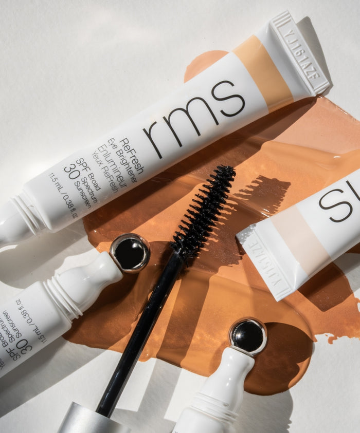 Close-up of RMS Beauty makeup products including an eye brightener with applicator and mascara wand, displayed on a neutral background with warm beige liquid makeup swatches, featuring The Wide Awake Duo.