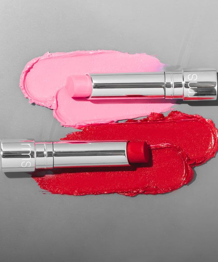Two RMS Beauty Tinted Daily Lip Balm sticks, one in soft pink and the other in vibrant red, displayed over matching creamy smears on a gray surface, showcasing clean beauty and natural ingredients.