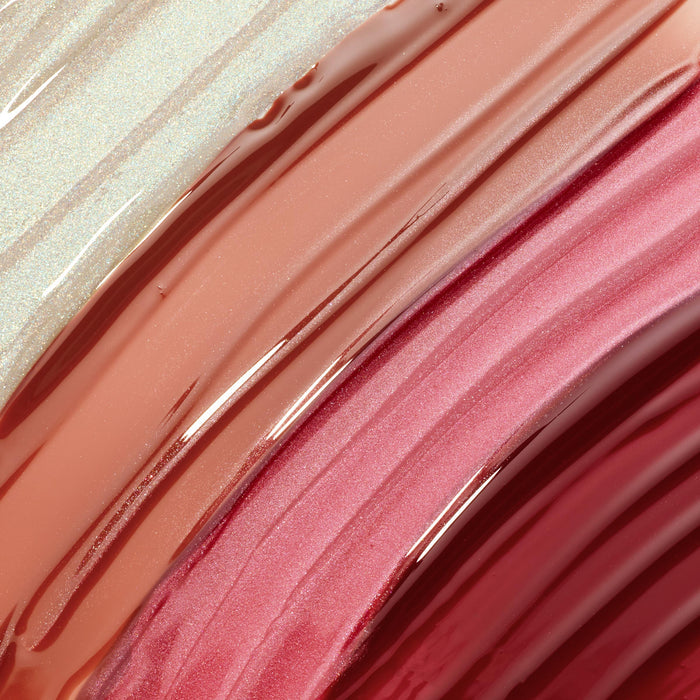 ColourPop Ultra Glossy Lip Totally Swamped swatch with companion shades, shown as thick diagonal streaks of high-shine gloss - champagne shimmer, peachy nude, and rosy pink - on a smooth surface to highlight texture and payoff.