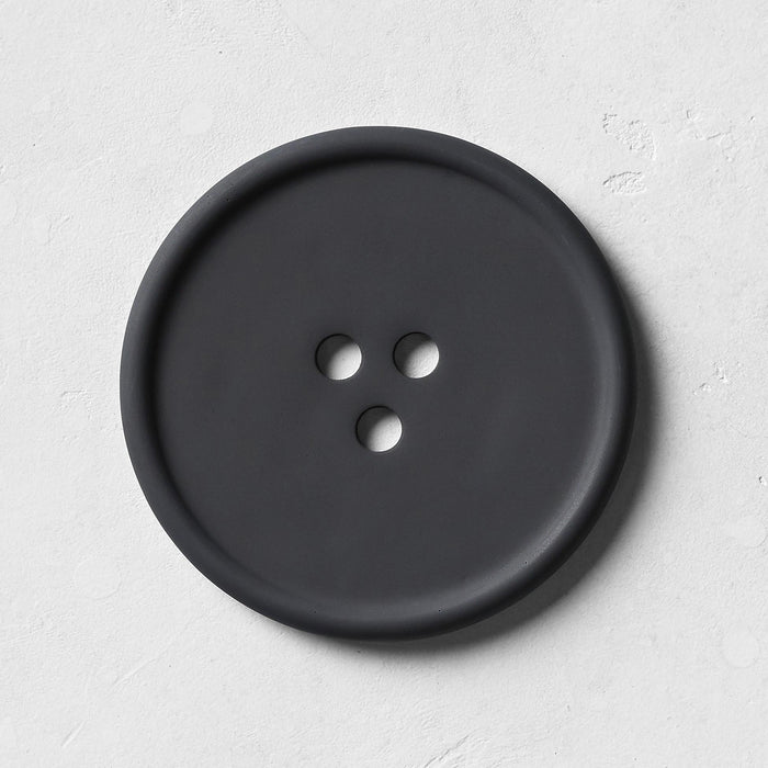 The UME Studio Black Bouton Erode Soap Dish, large and round with three holes, rests on a handmade light gray textured coaster, casting a soft shadow to the right.