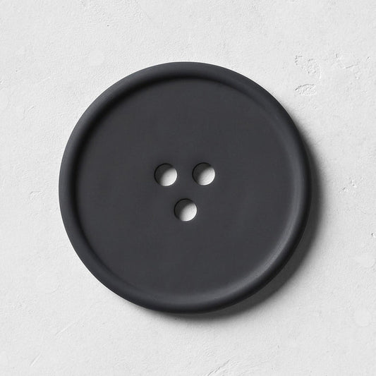 The UME Studio Black Bouton Erode Soap Dish, large and round with three holes, rests on a handmade light gray textured coaster, casting a soft shadow to the right.