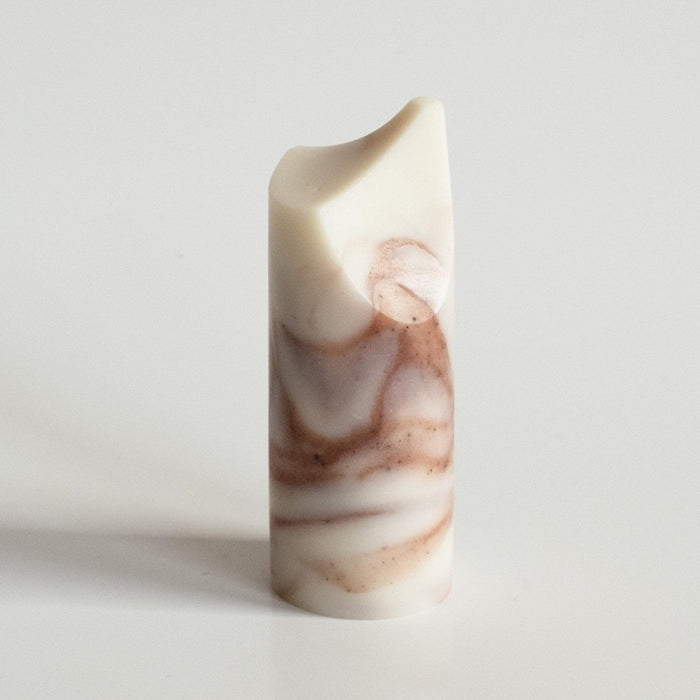 The UME - LAVENDER CREME MINI ERODE SOAP by UME STUDIO is a cylindrical bar with marbled white and brown swirls, standing upright on a white surface. Its top edge is slightly curved and uneven.