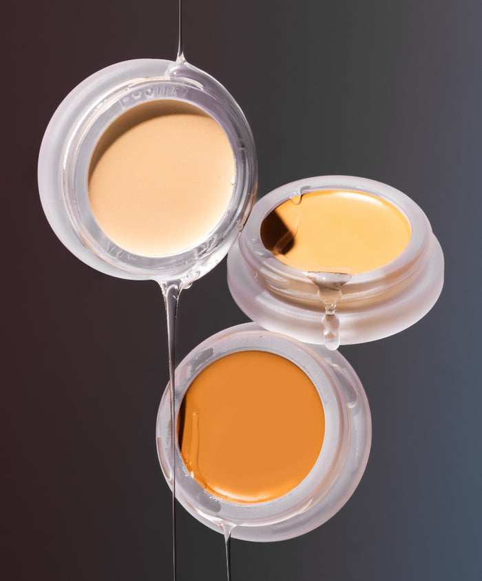 Three open jars of UnCoverup Concealer in varying shades with a clear liquid drizzling over them, highlighting the smooth, creamy texture and natural ingredients in clean beauty packaging.