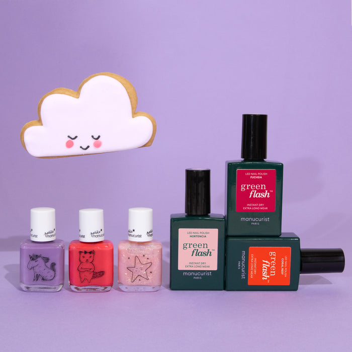 Manucurist's You  Me at a Tea Party Green Flash™ Set is displayed on a lavender background with a smiling cloud-shaped cookie nearby. Some bottles feature animal and star illustrations.