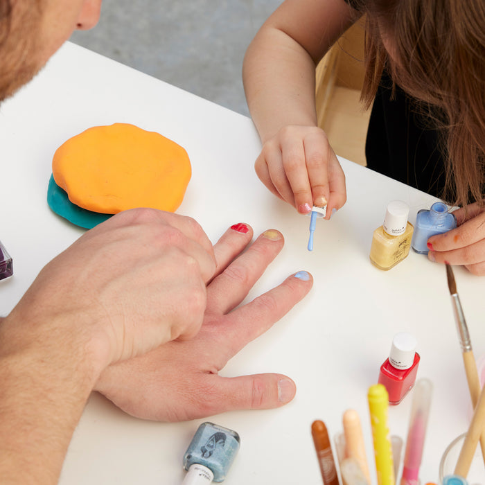 A child uses the Manucurist You  Me at a Tea Party Green Flash™ Set to paint an adult’s nails blue at a white table, surrounded by colorful nail polish bottles, markers, and orange playdough.