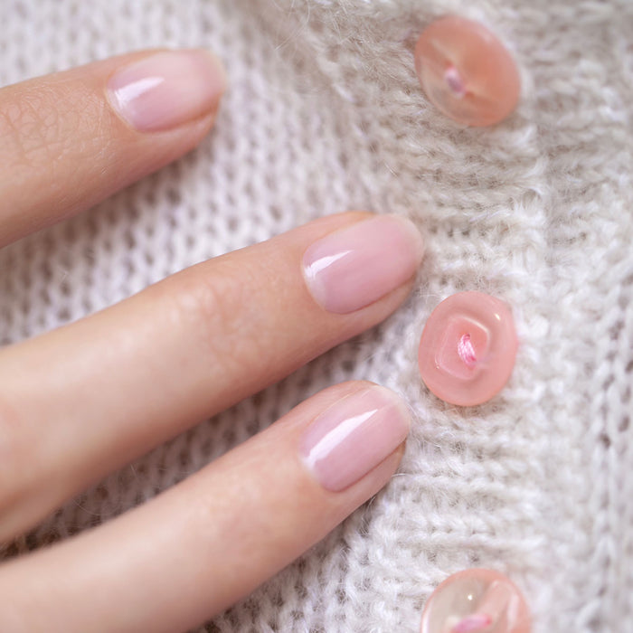 A close-up of a hand with glossy, neatly manicured light pink nails—painted with the Manucurist You  Me at a Tea Party Green Flash™ Set—resting on white knit fabric adorned with pink translucent buttons.