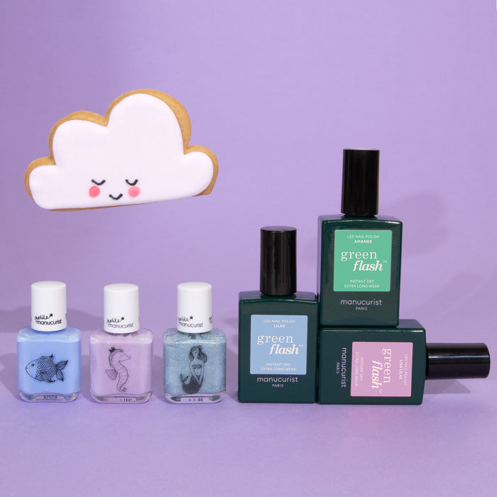 Five bottles from the Manucurist You  Me Under the Sea Green Flash™ Set are arranged on a purple surface. Three pastel shades with illustrated animal labels are featured, with a smiling cloud-shaped cookie in the background.
