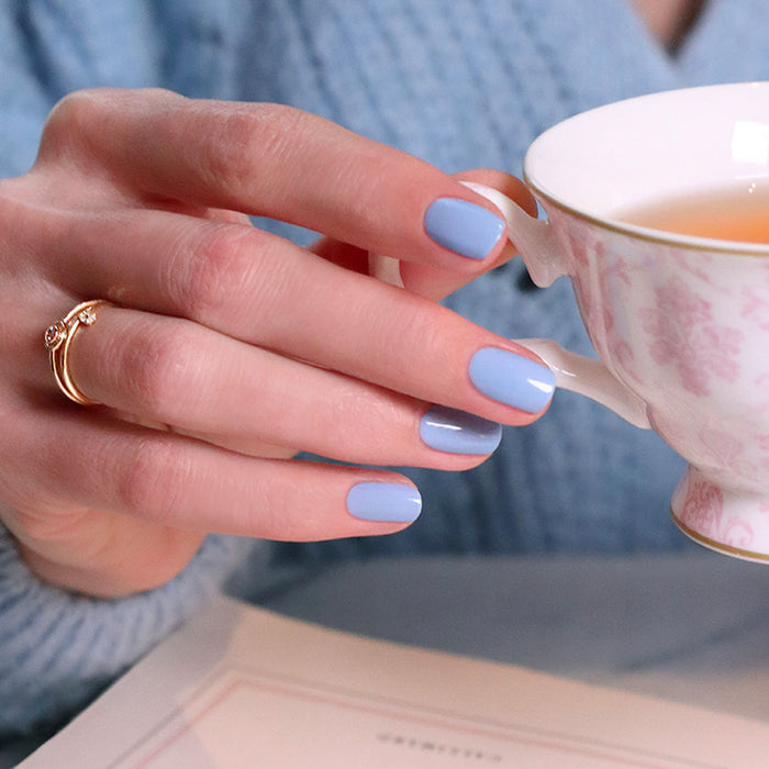 A hand with neatly manicured nails polished in Manucurist's You  Me Under the Sea Green Flash™ Set holds a white teacup with a pink floral design. The person wears a gold ring, blue sweater, and an open book is partially visible.