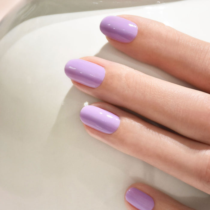 A close-up of a hand with polished nails painted in glossy lavender from the Manucurist You  Me Under the Sea Green Flash™ Set, resting on a smooth white surface.