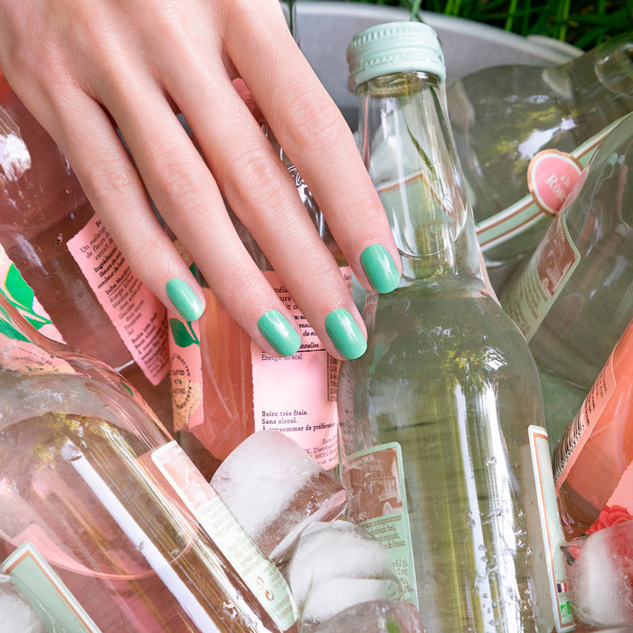 A hand with Manucurist's You  Me Under the Sea Green Set nail polish rests on glass bottles of pink and clear drinks, surrounded by ice cubes in a metal container.